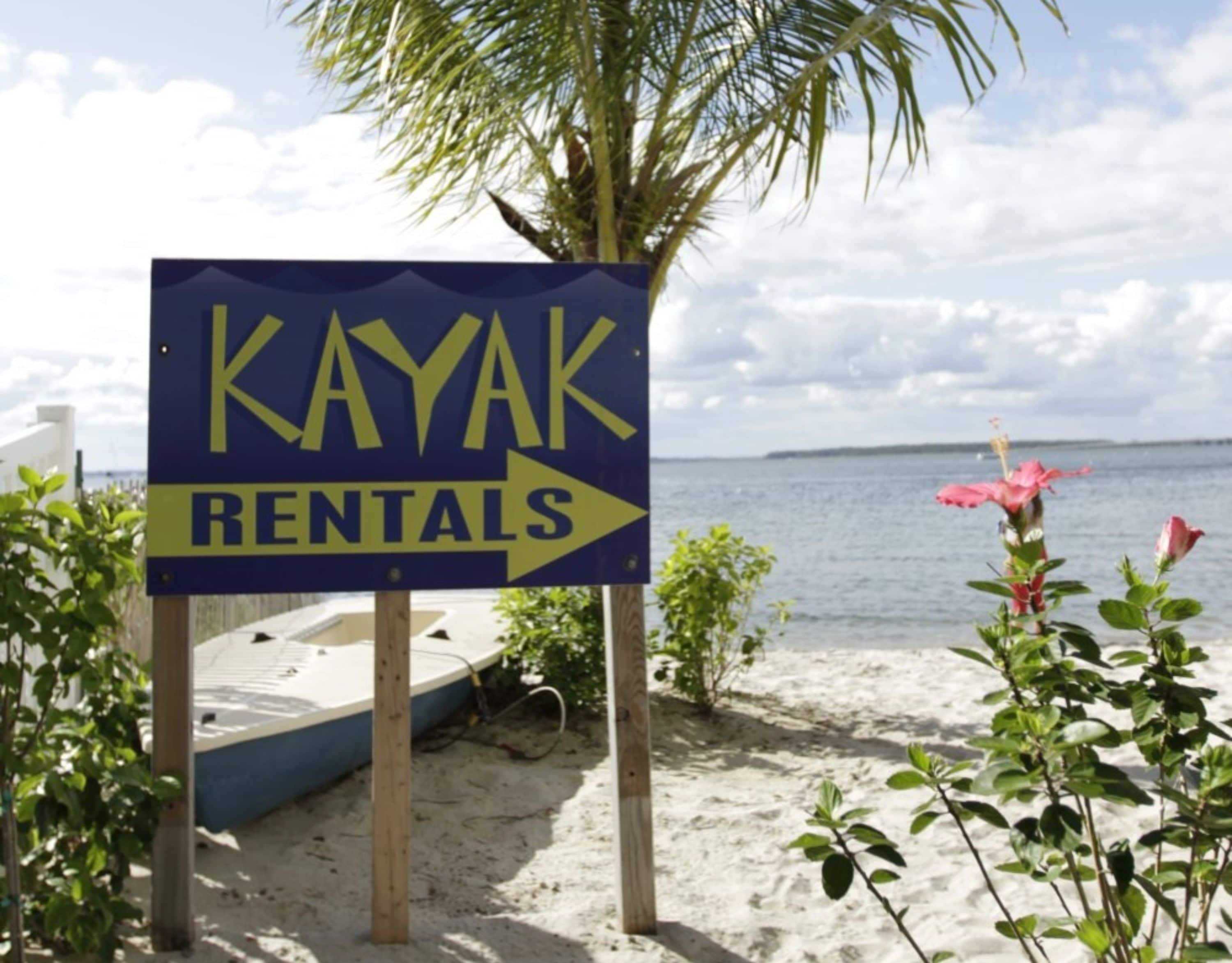 Señal para alquiler de kayaks con una flecha apuntando a la derecha, ubicada en una playa arenosa con una masa de agua al fondo.