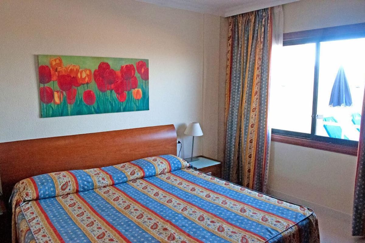Ein Hotelzimmer mit einem Bett, einem Bild von roten Blumen an der Wand, einem Fenster mit Vorhängen und Blick ins Freie.