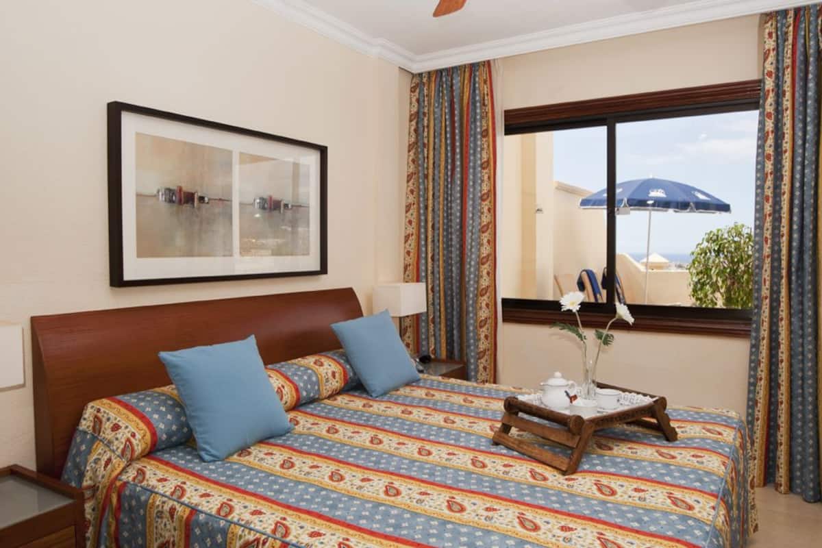 Ein Hotelzimmer mit einem großen Bett, einem gerahmten Bild an der Wand, einem Fenster mit Vorhängen und einem kleinen Tisch mit Tablett.