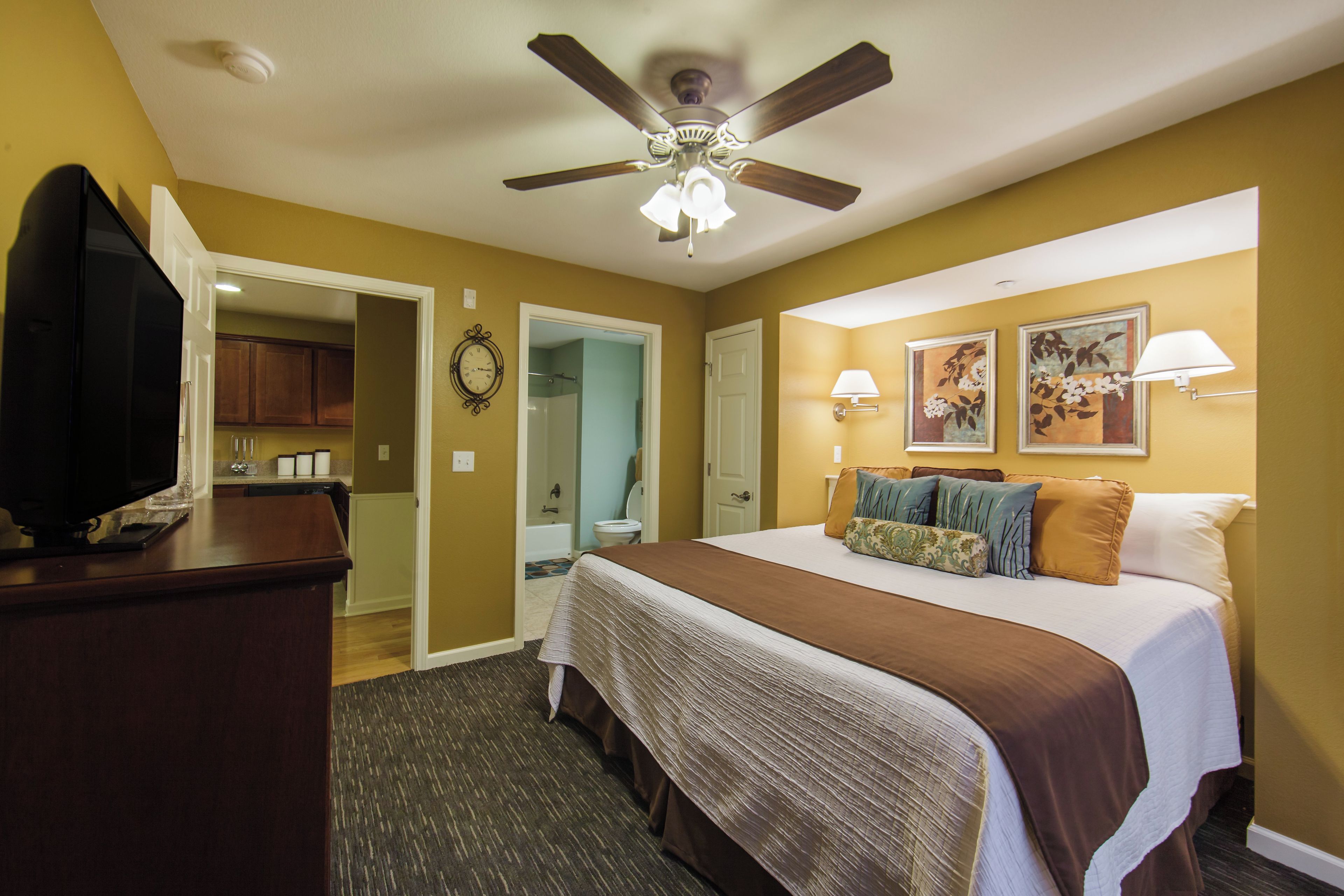 A hotel room with a bed, a television on a stand, a ceiling fan, a bathroom with a shower, and a hallway leading to a kitchen.