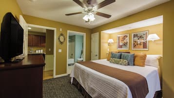 A hotel room with a bed, a television on a stand, a ceiling fan, a bathroom with a shower, and a hallway leading to a kitchen.