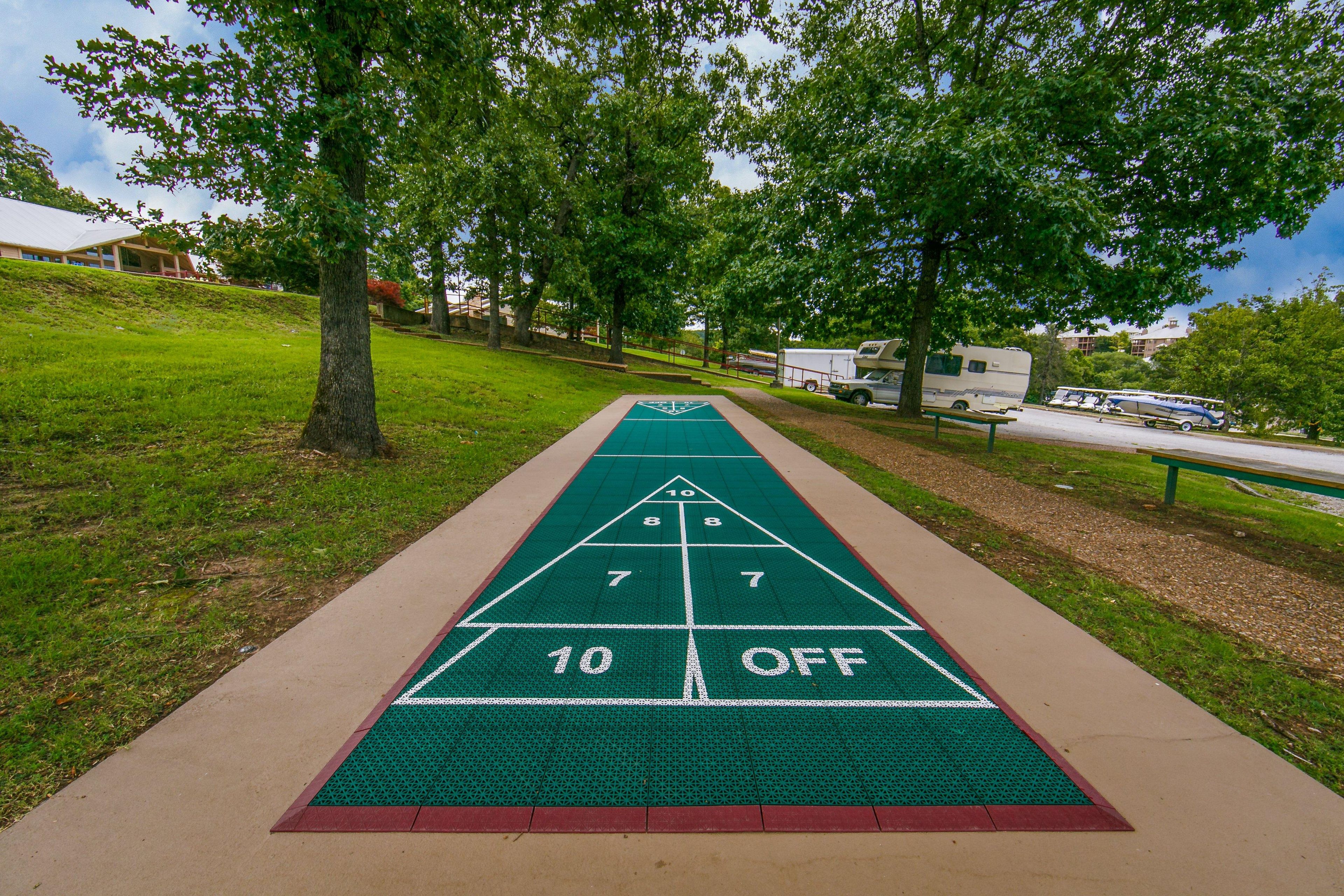 Una cancha de shuffleboard con superficie verde y marcas blancas, rodeada de césped y árboles.