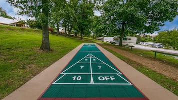 Una cancha de shuffleboard con superficie verde y marcas blancas, rodeada de césped y árboles.