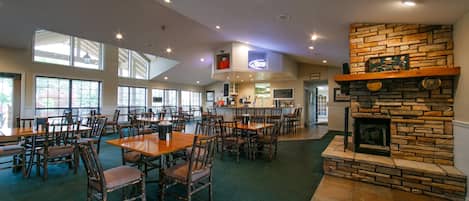 A spacious dining area with multiple tables and chairs, a stone fireplace, and large windows providing natural light.