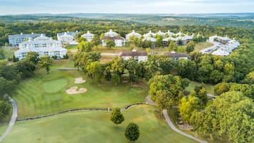 Vue aérienne d’un terrain de golf avec plusieurs bâtiments, dont un club-house et des logements, le tout entouré d’une végétation luxuriante.