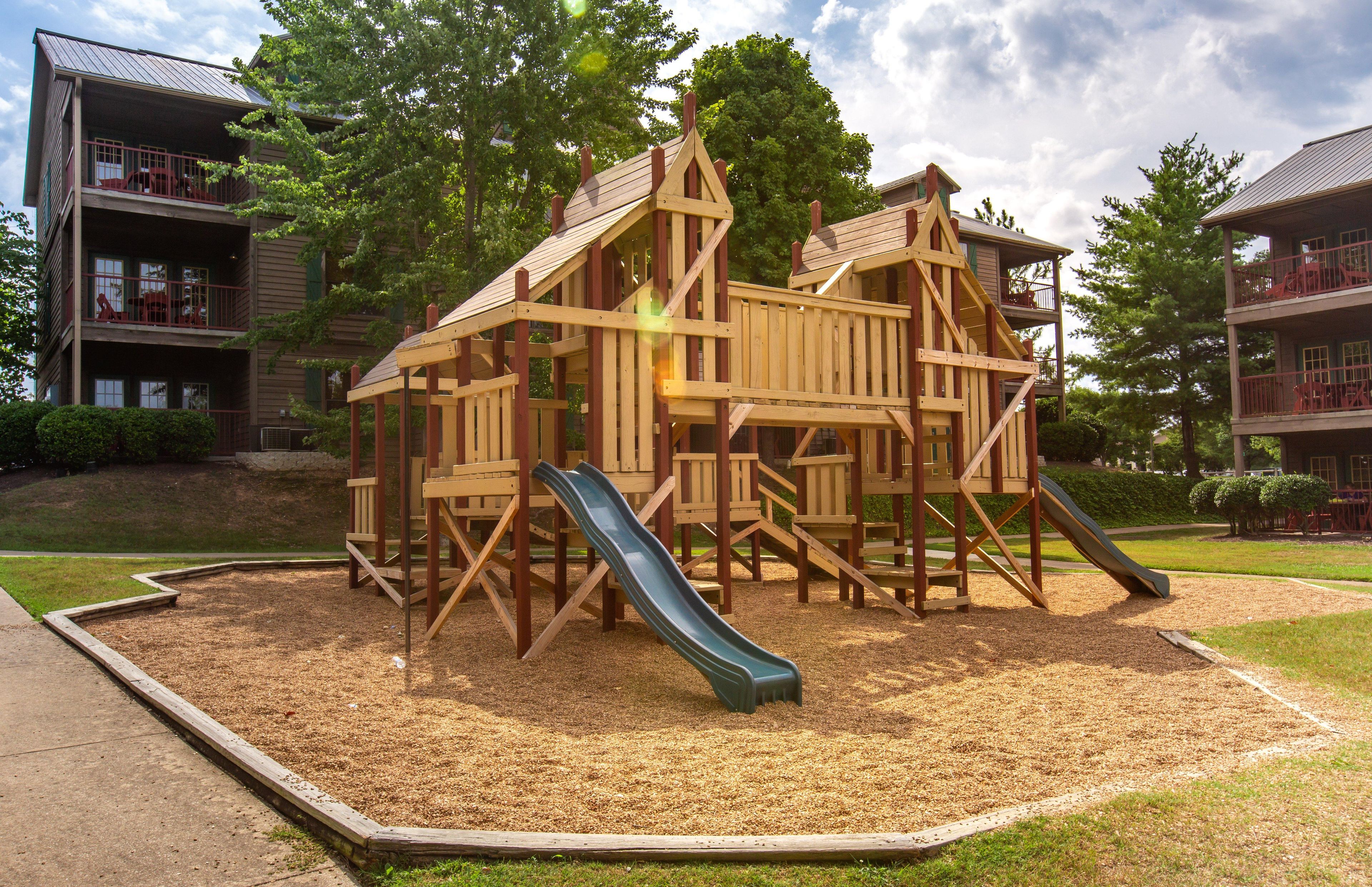 Un parc infantil amb tobogan i elements per escalar, envoltat d'una zona verda i arbres.