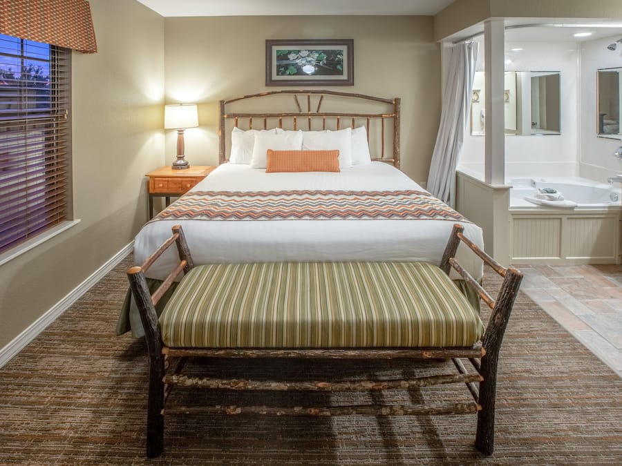 Habitación de hotel con una cama grande, una silla de madera, una mesita de noche con lámpara, un baño con bañera y una ventana con persianas.