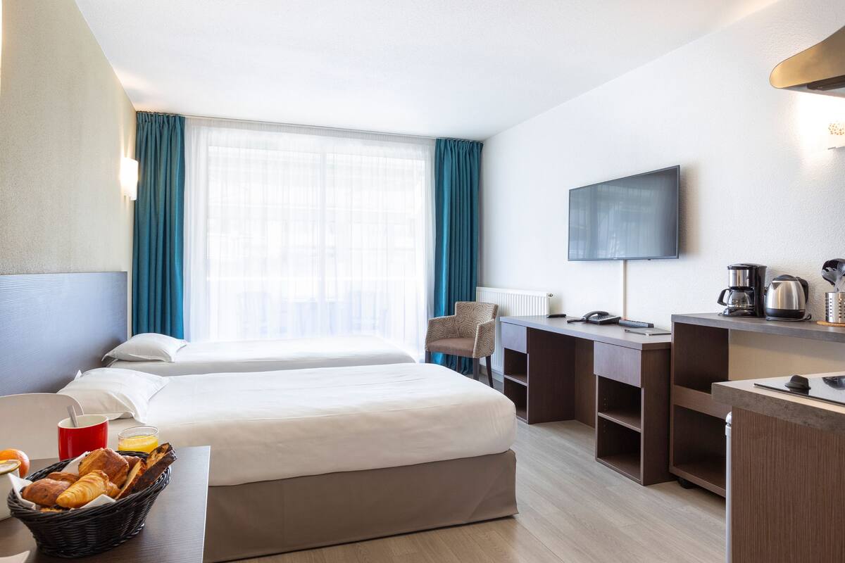 Ein Hotelzimmer mit zwei Betten, einem Schreibtisch, einem Fernseher, einer Kaffeemaschine und einem Frühstückswagen mit Brot und Obst.