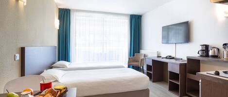 A hotel room with two beds, a desk, a TV, a coffee maker, and a breakfast tray with bread and fruit.