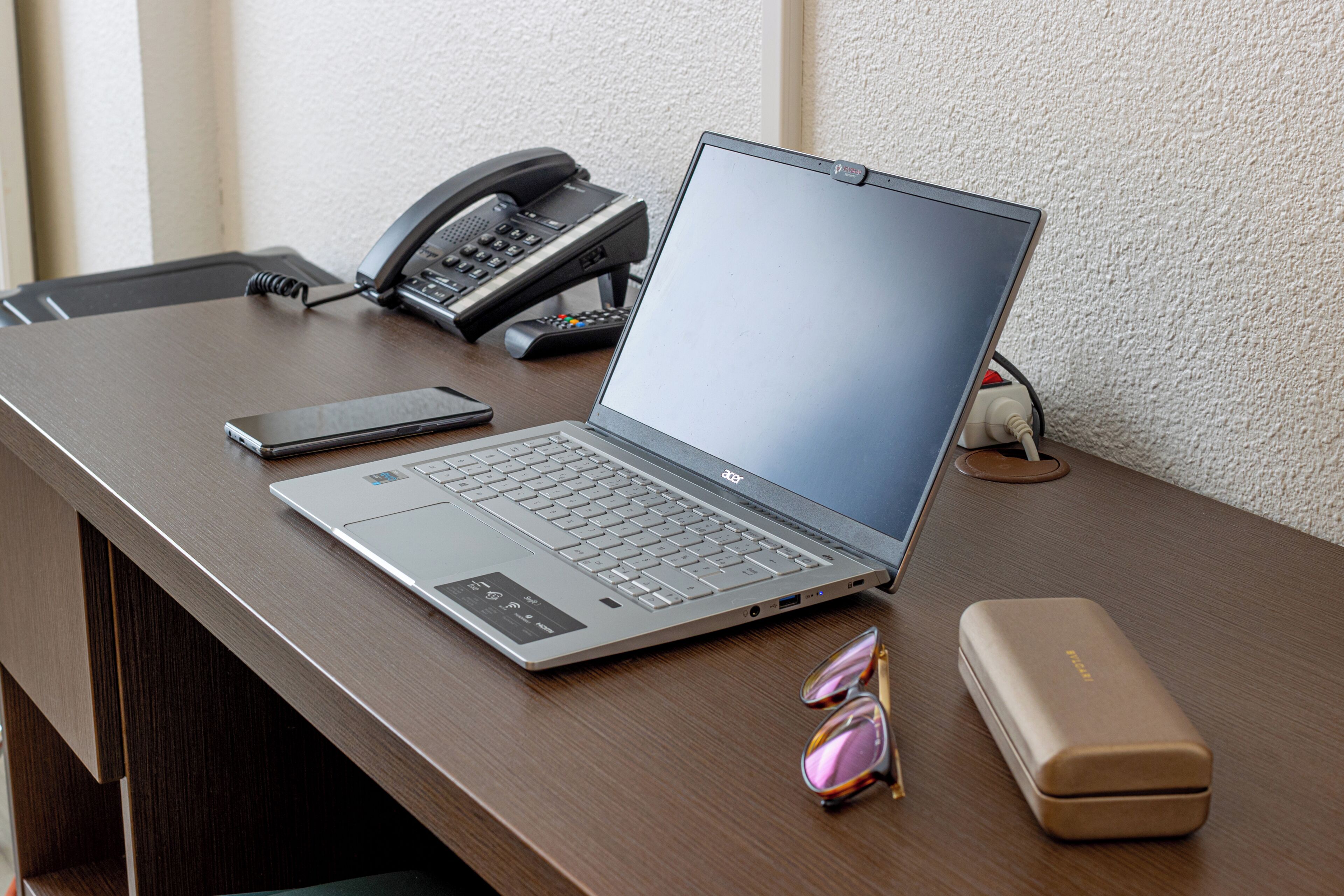 A laptop on a desk with a phone, glasses, and a charging cable.