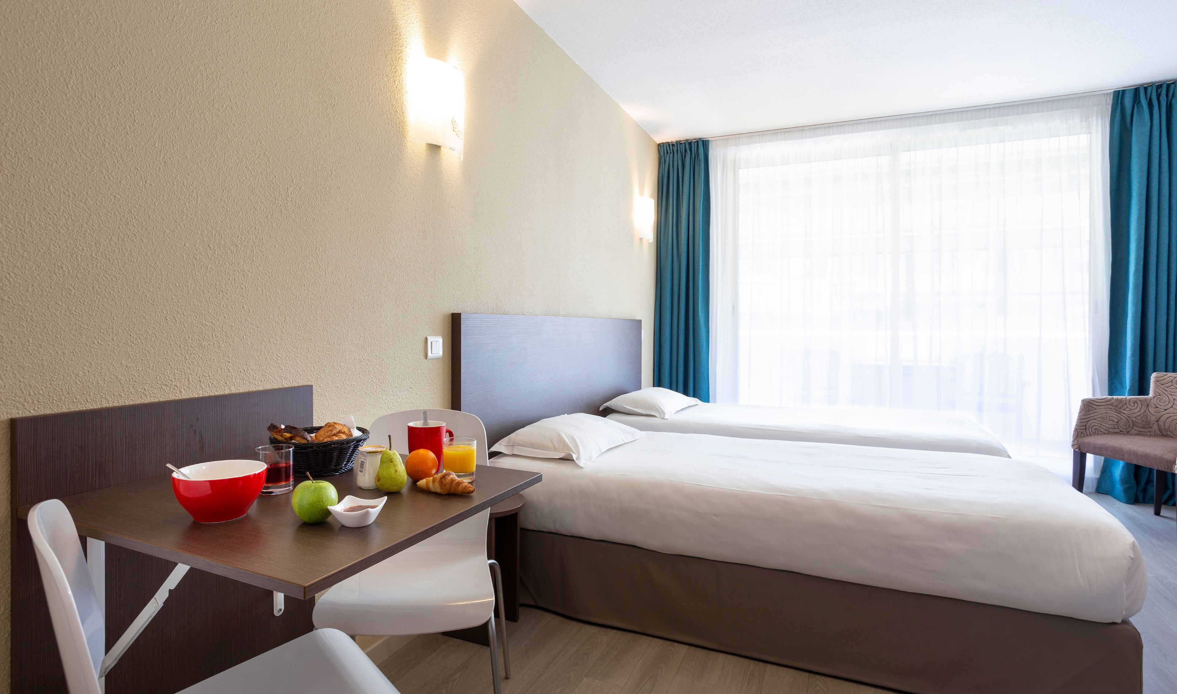 Une chambre d’hôtel avec un lit, un bureau avec des articles pour le petit-déjeuner, une chaise et une fenêtre avec des rideaux.