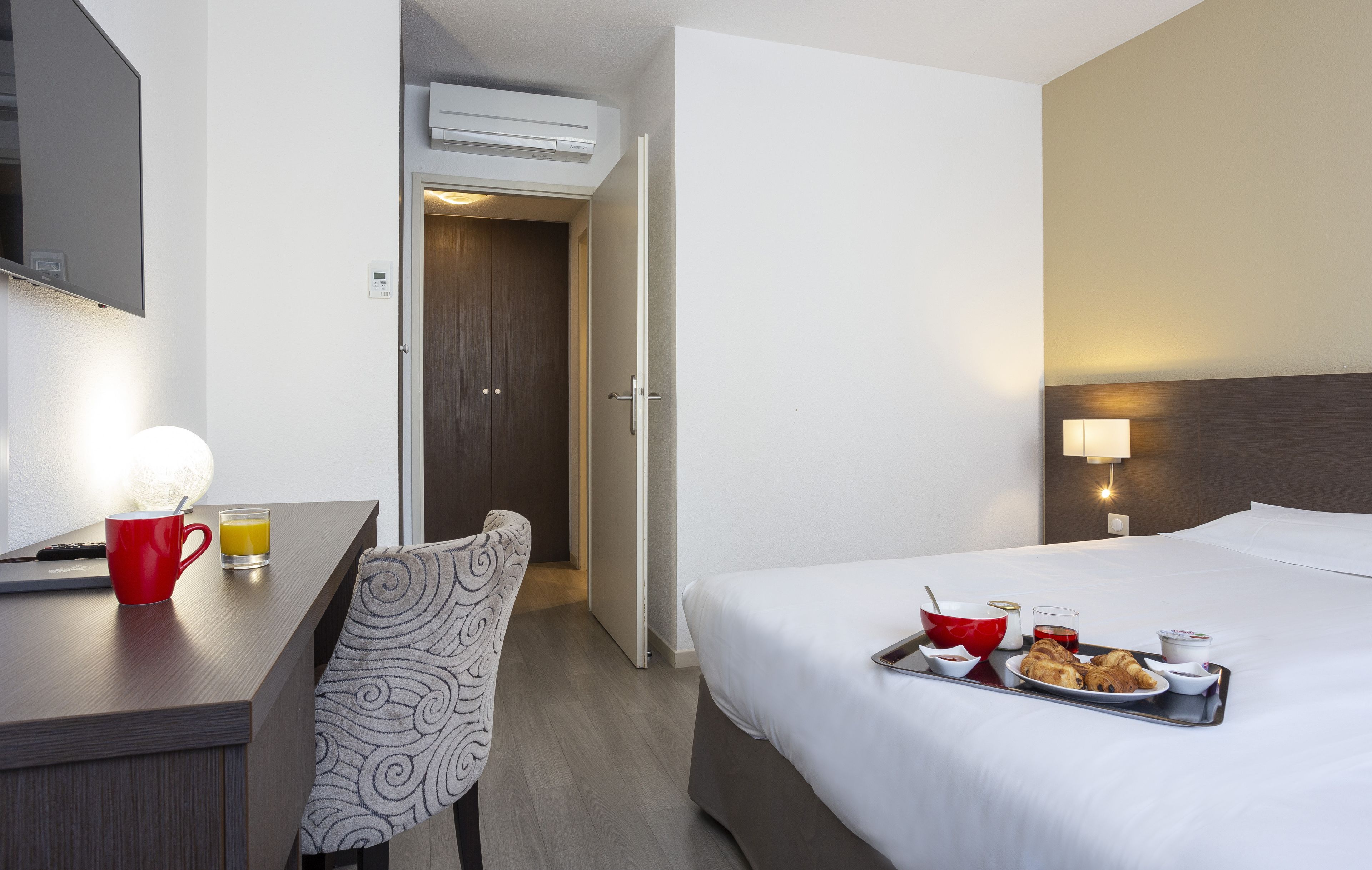 Una habitación de hotel con cama, mesita de noche, silla y bandeja de desayuno con croissants y bebidas.