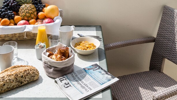 A balcony with breakfast items, a newspaper, and a view of a building and trees. - Les Loges du Park – résidence hôtelière (Aix-les-Bains)