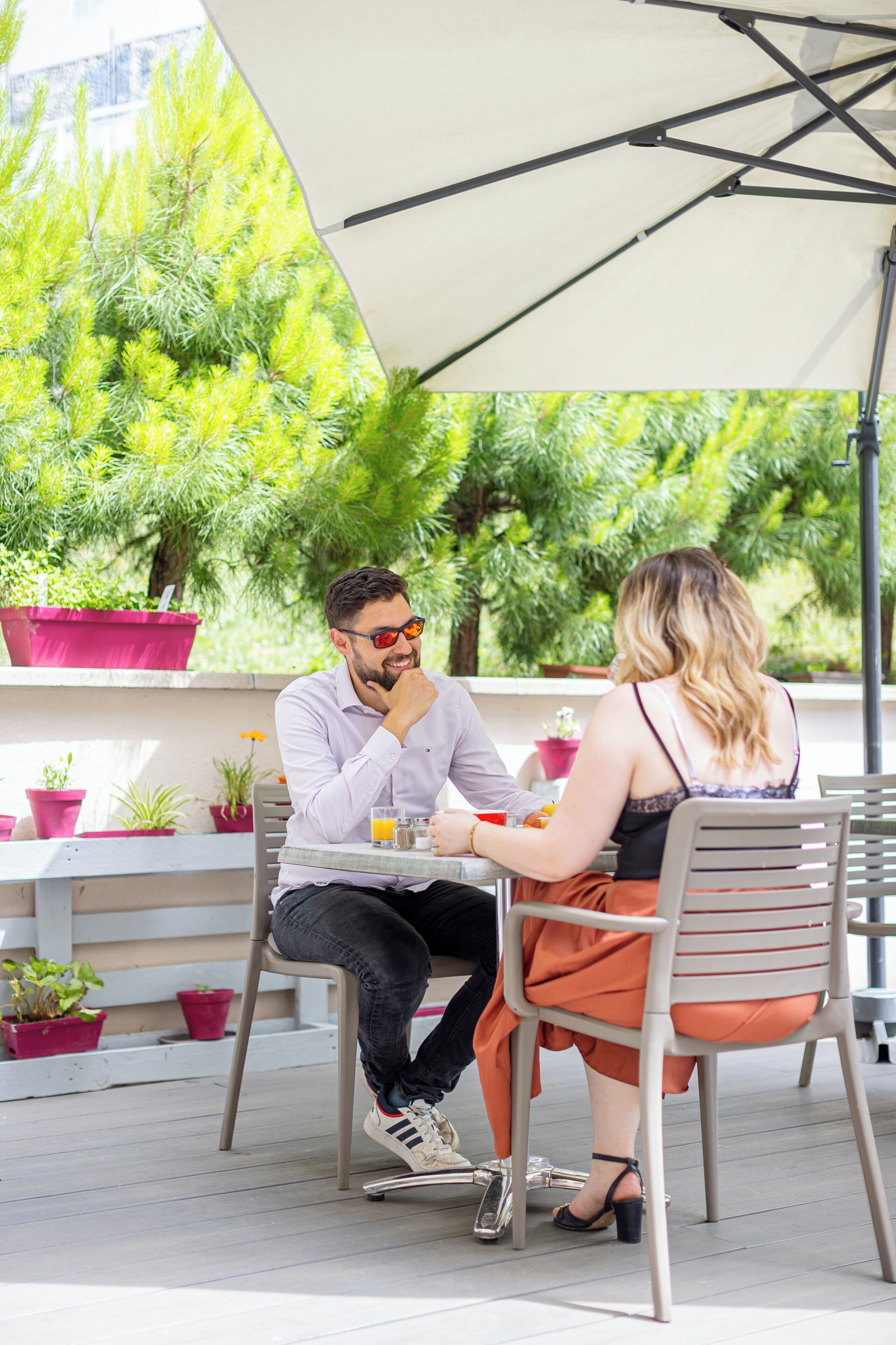 Dos personas sentadas en una mesa al aire libre con una sombrilla blanca, rodeadas de plantas en macetas y follaje.