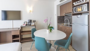 A compact kitchen with a microwave, refrigerator, and sink. There's a dining table with chairs and a potted orchid. A flat-screen TV is mounted on the wall. - Les Loges du Park – résidence hôtelière (Aix-les-Bains)