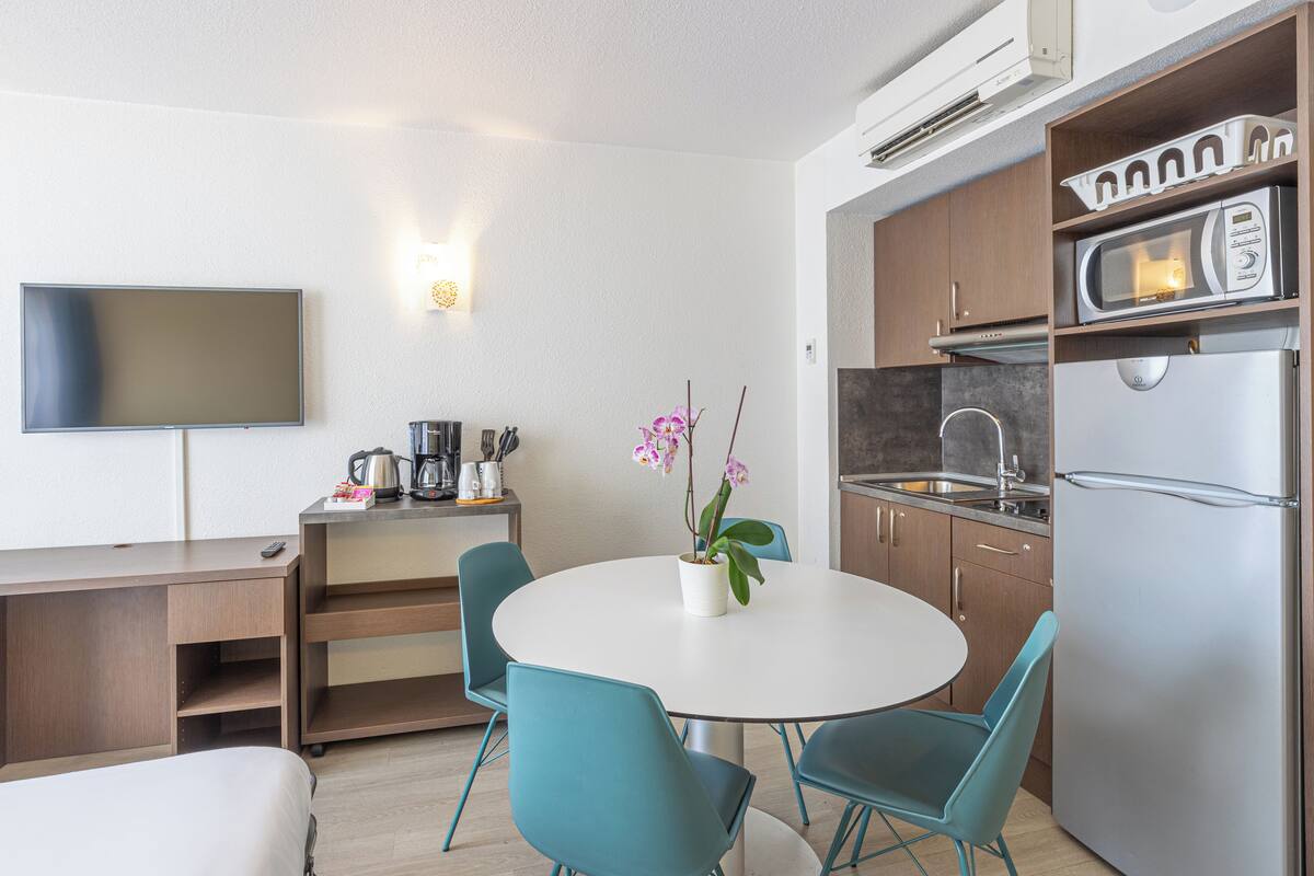 Eine kompakte Küche mit Mikrowelle, Kühlschrank und Spüle. Ein Esstisch mit Stühlen und ein Topfhanf-Orchideen stehen bereit. Ein Flachbildfernseher ist an der Wand befestigt.