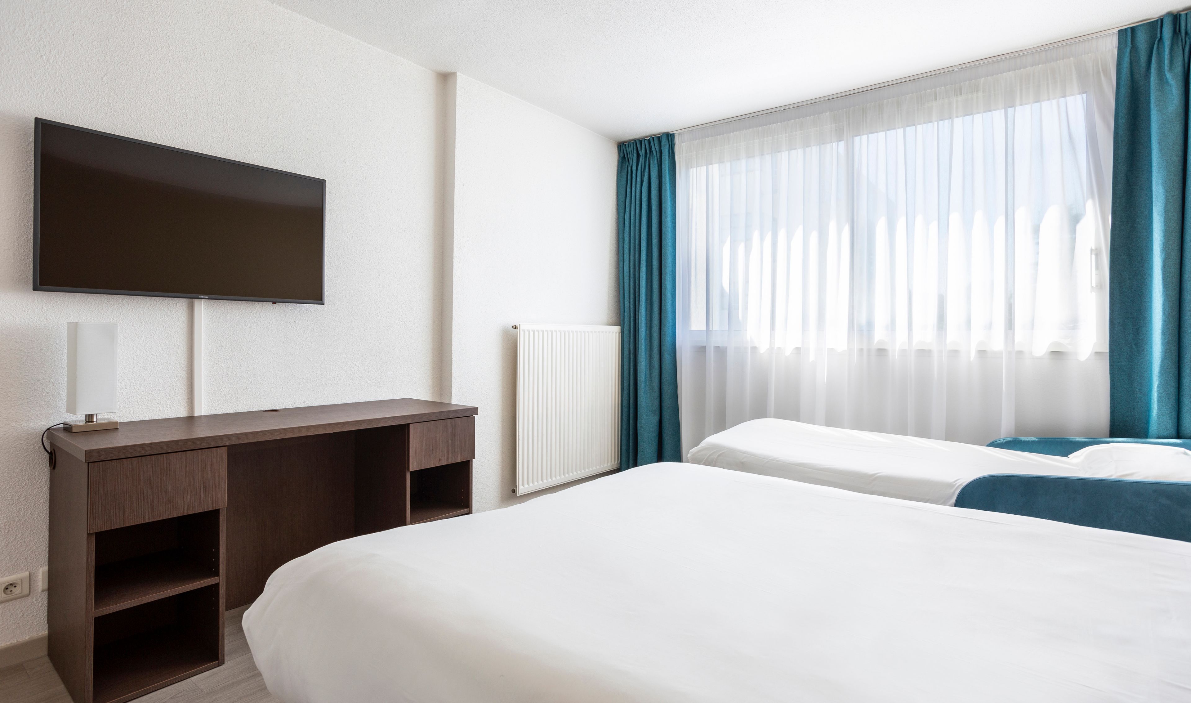 A hotel room with a bed, a desk with a lamp, a television, and a window with curtains.
