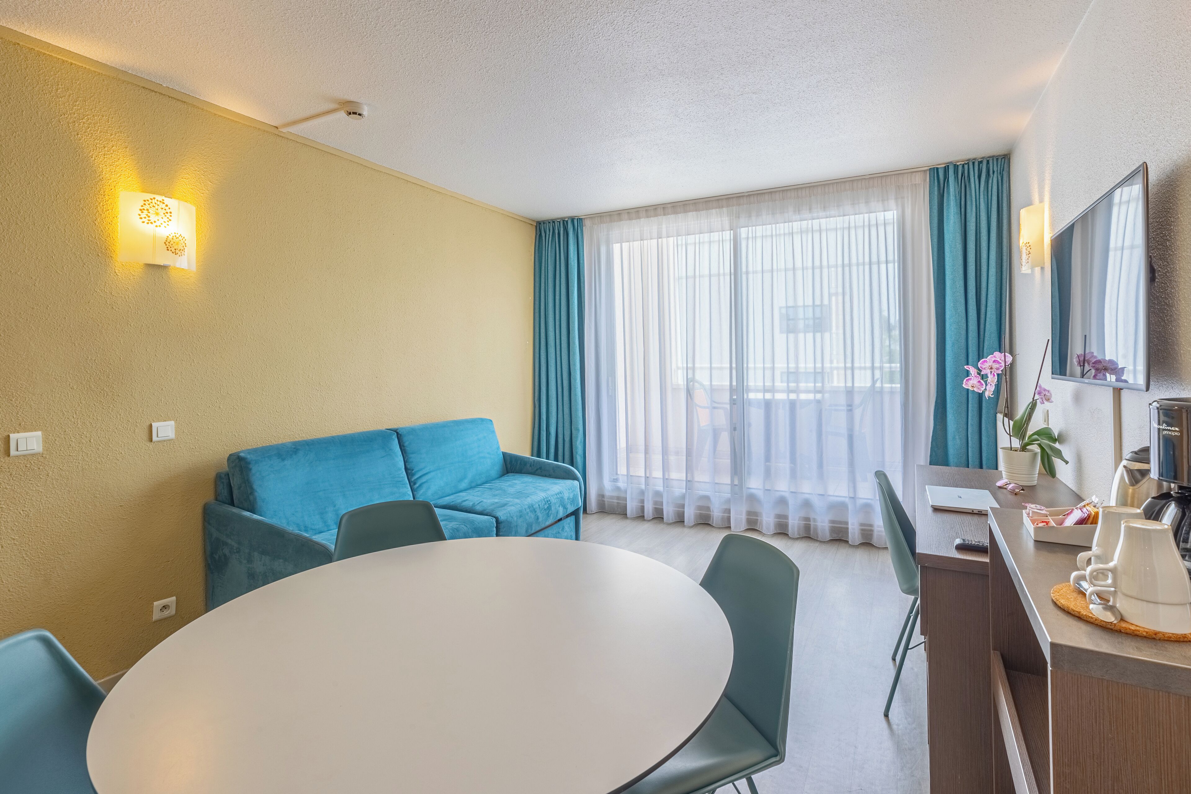 A hotel room with a round dining table, a blue sofa, a TV, and a window with curtains.