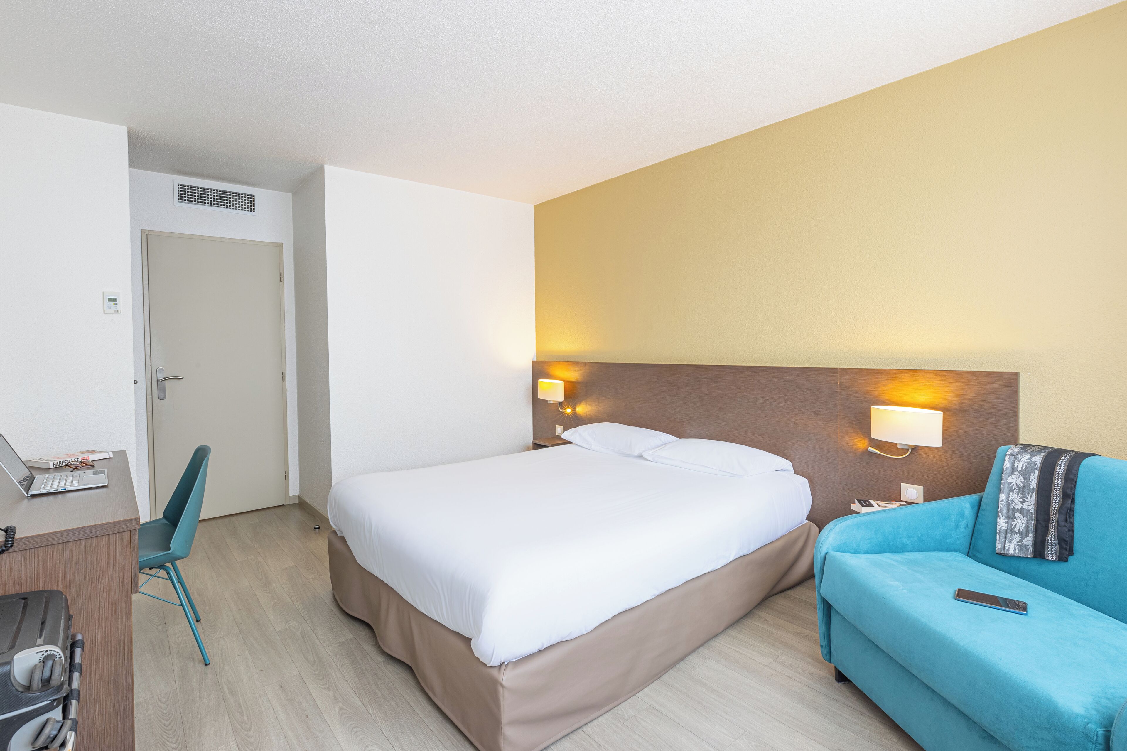 Een hotelkamer met een bed, een bureau met een laptop, een stoel, een bank en twee wandlampen.