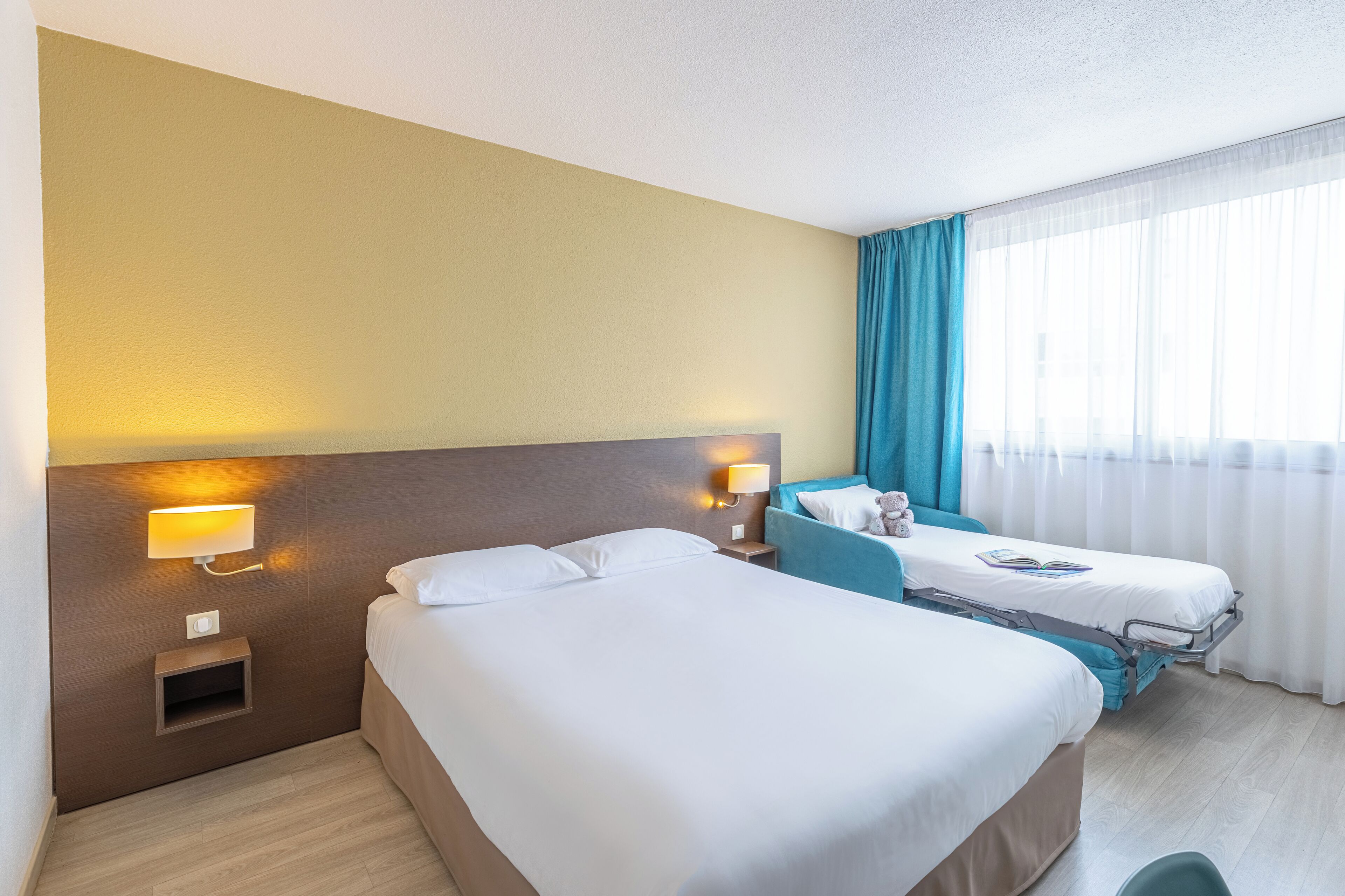 Ein Hotelzimmer mit zwei Betten, einem Nachttisch mit Lampe, einem kleinen Tisch und einem Fenster mit Vorhängen.