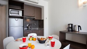 A compact kitchen with a microwave, sink, and coffee maker. A breakfast table with fruits, pastries, and coffee cups is set. - Les Loges du Park – résidence hôtelière (Aix-les-Bains)