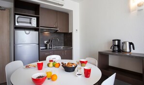 Une cuisine compacte équipée d’un four à micro-ondes, d’un évier et d’une machine à café. Une table de petit-déjeuner est dressée, avec des fruits, des viennoiseries et des tasses à café.