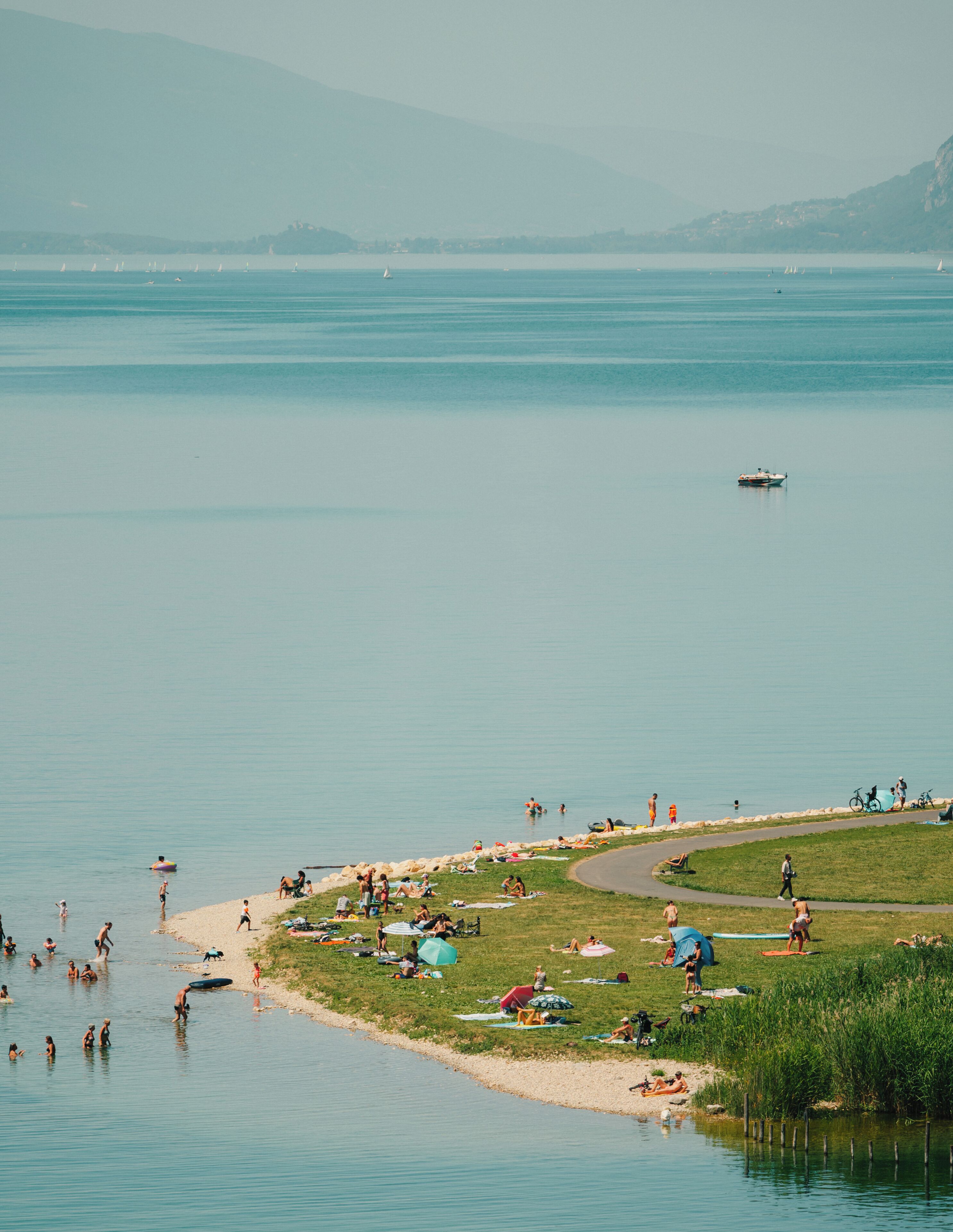 Une vue sur le lac, avec des personnes qui se baignent et prennent des bains de soleil, un bateau et une zone herbeuse où l’on peut faire du vélo.