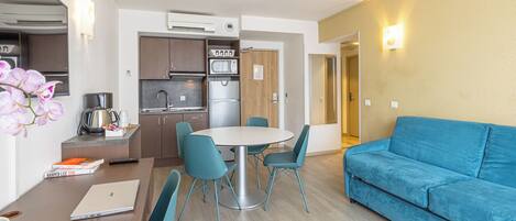 A compact living space with a dining table, chairs, a sofa, a kitchen area, and a wall-mounted light fixture.
