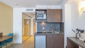 A compact kitchen with a microwave, refrigerator, and sink. There is a dining table with chairs and a wooden door leading to another room. - Les Loges du Park – résidence hôtelière (Aix-les-Bains)