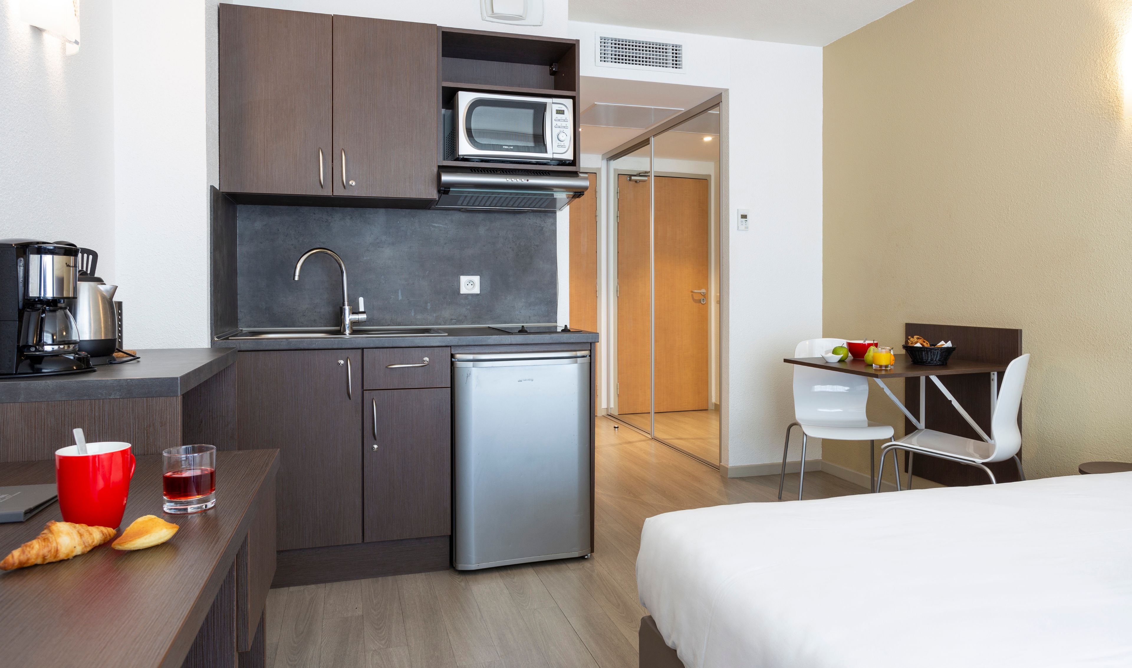 A compact hotel room with a kitchenette, a bed, a small desk, and a chair.