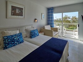 Habitación de hotel con dos camas, un balcón con sillas blancas y vista a zonas verdes.