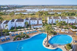 Ein Resort mit mehreren weißen Villen, einem zentral gelegenen Pool und Blick auf eine natürliche Landschaft mit Gewässern und Palmen.