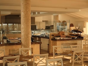 A modern hotel dining area with a central island, seating, and a view of the kitchen. - Viva Alcudia Sun Village (Muro)