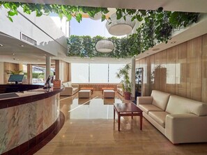 A modern hotel lobby with a reception desk, seating area, and large windows. - Viva Alcudia Sun Village (Muro)