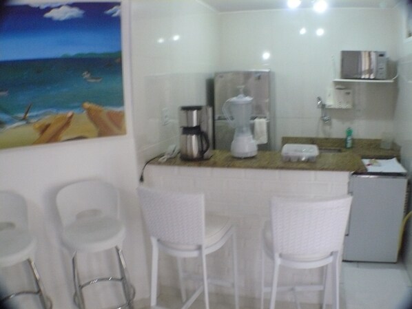 A small kitchenette with a coffee maker, blender, and microwave. There are three white chairs and a small refrigerator.