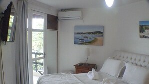 A hotel room with a bed, a painting of a coastal scene, an air conditioning unit, and a view through a door. - BÚZIOS BEACH INTERNACIONAL RESIDENCE SERVICE (Búzios)