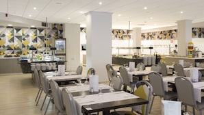 A modern dining area with multiple tables and chairs, a kitchen area in the background, and geometric patterned walls. - Aparthotel Acuasol (Peniscola)