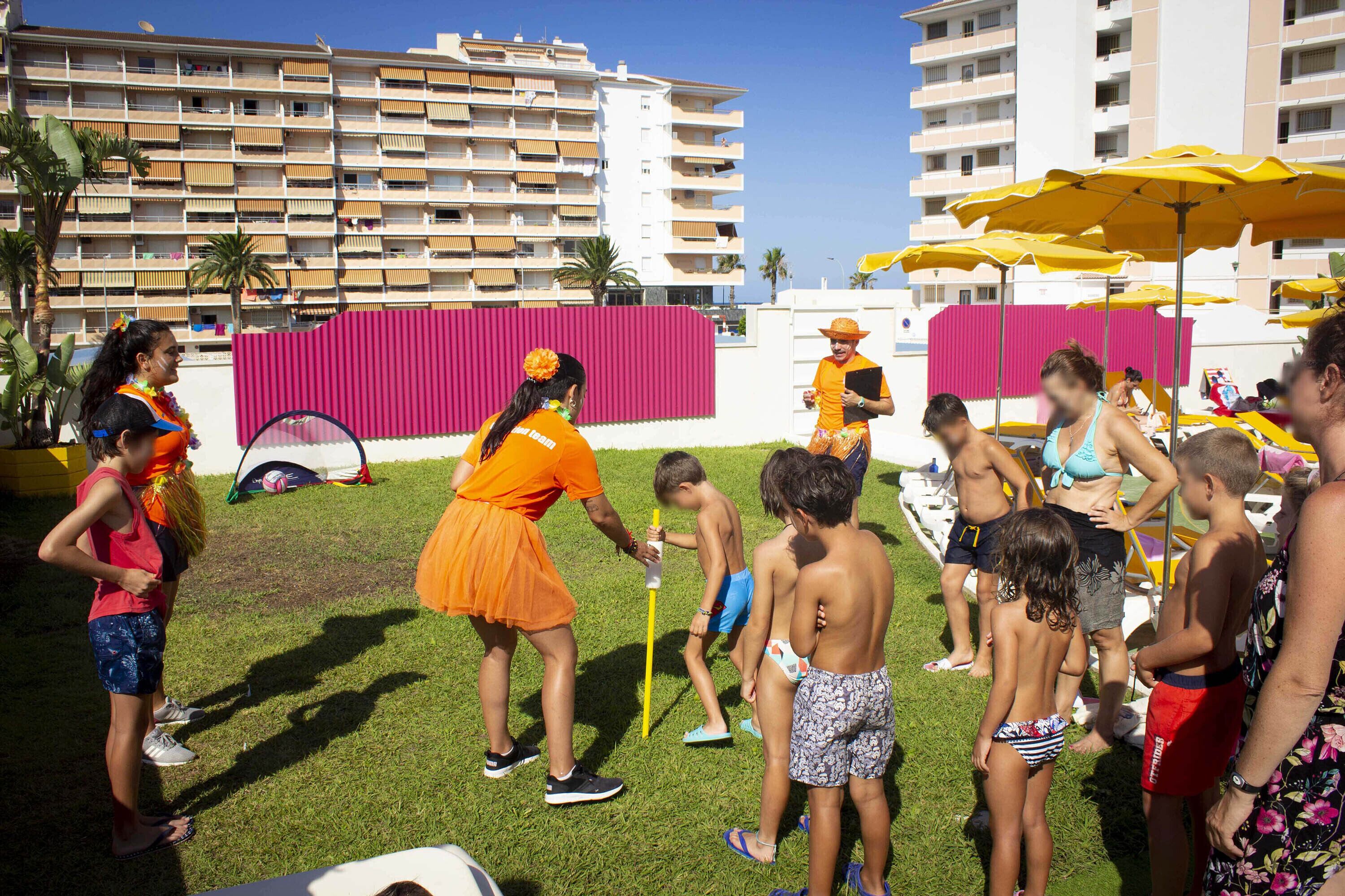 Un grup de persones reunides a l'aire lliure, possiblement en un centre turístic o zona de platja, amb edificis i paraigües al fons.