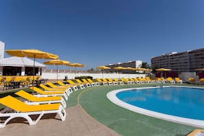 Zona de piscina con tumbonas amarillas y sombrillas, un bar y un edificio al fondo.