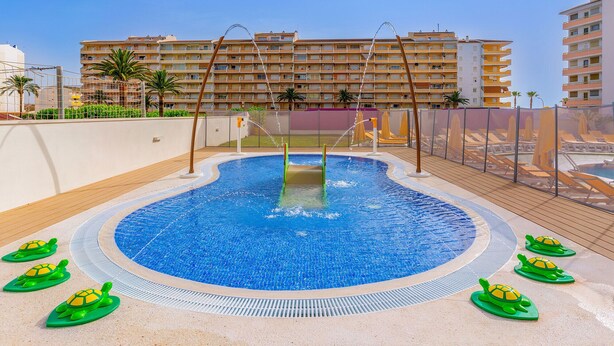 A swimming pool with a slide, surrounded by a deck area with sun loungers and a view of a multi-story building in the background. - Aparthotel Acuasol (Peniscola)