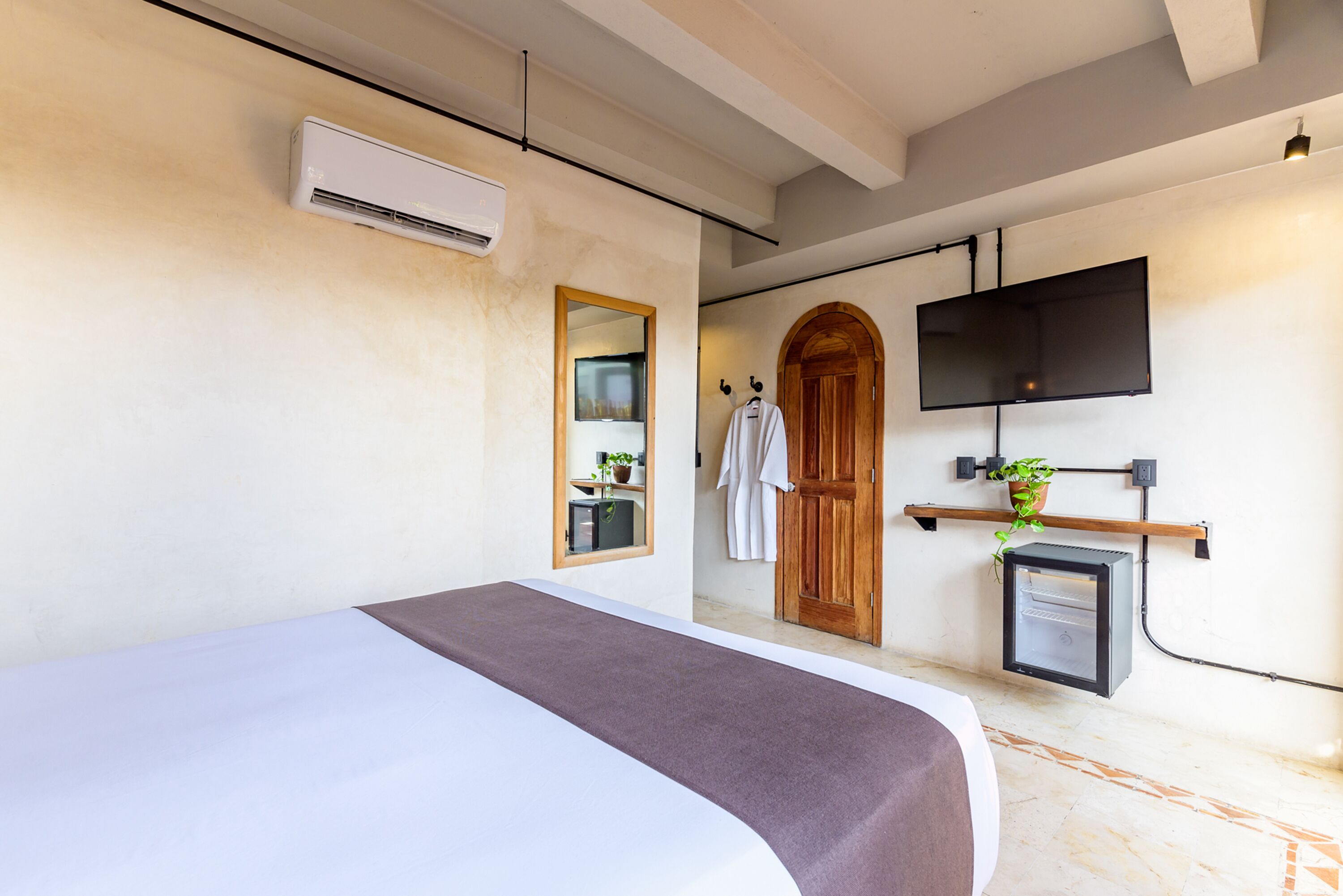 A hotel room with a bed, an air conditioning unit, a television mounted on the wall, a wooden door, and a small plant on a shelf.