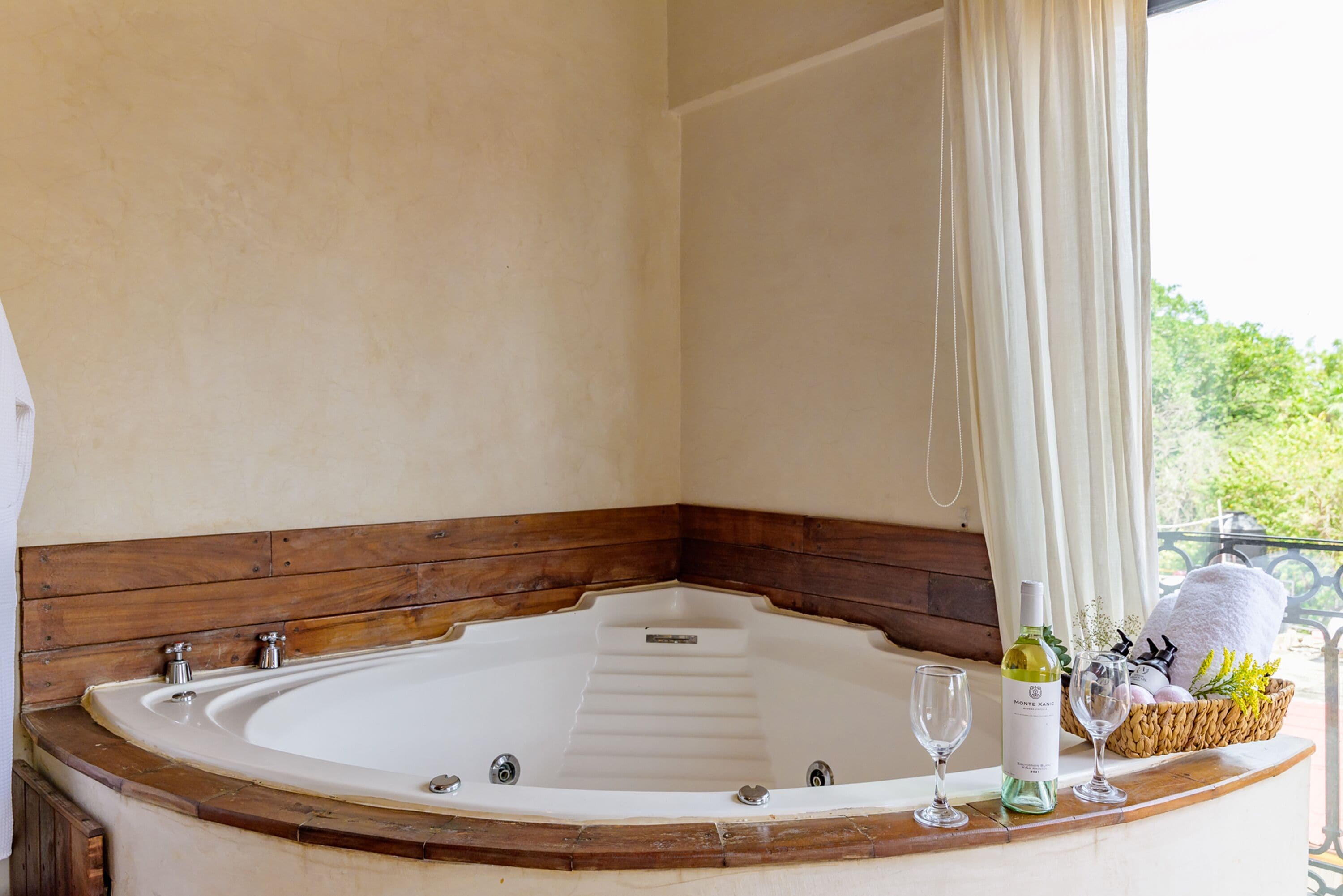 A corner bathtub with wooden trim, a bottle of wine, and two glasses on a tray.