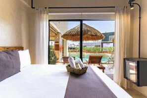 Habitación de hotel con cama, una mesita con toallas y botellas, un paraguas de paja y vista a un área de piscina al aire libre.