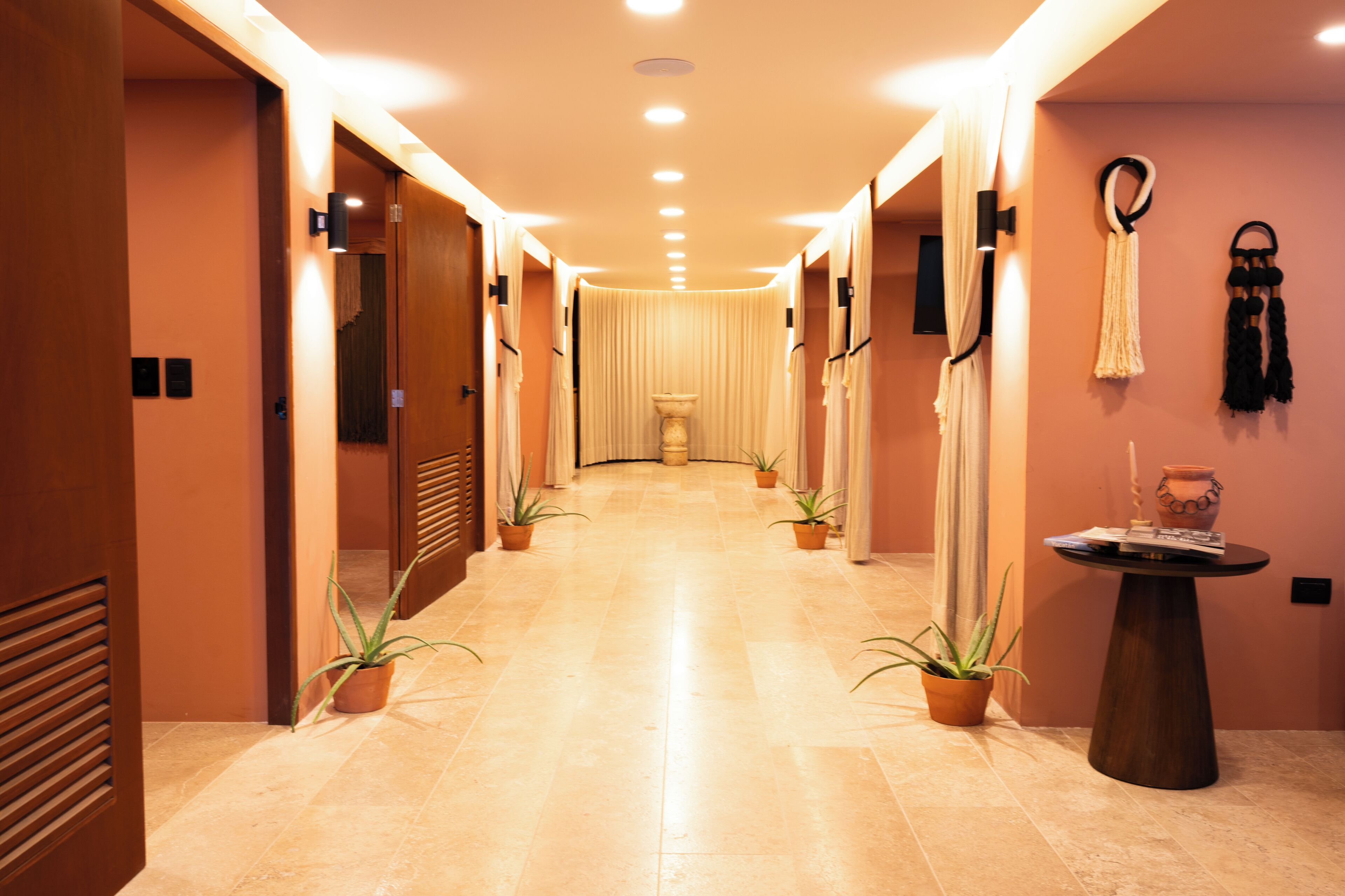 Un couloir d’hôtel avec un sol carrelé, des plantes en pot, une table ronde surmontée d’un vase et un téléviseur fixé au mur.