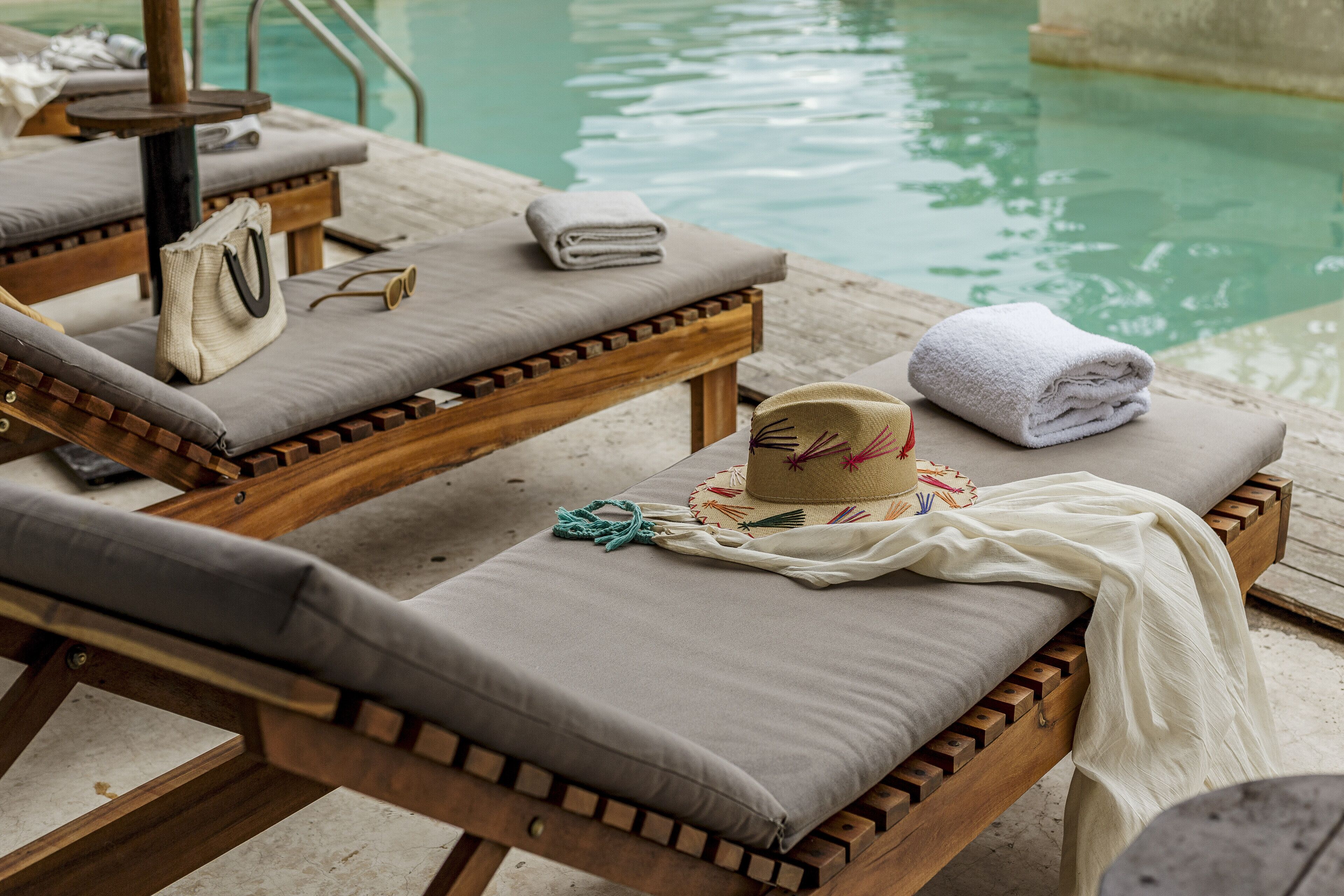 A wooden lounge chair with a cushion, a hat, and a towel by a pool.