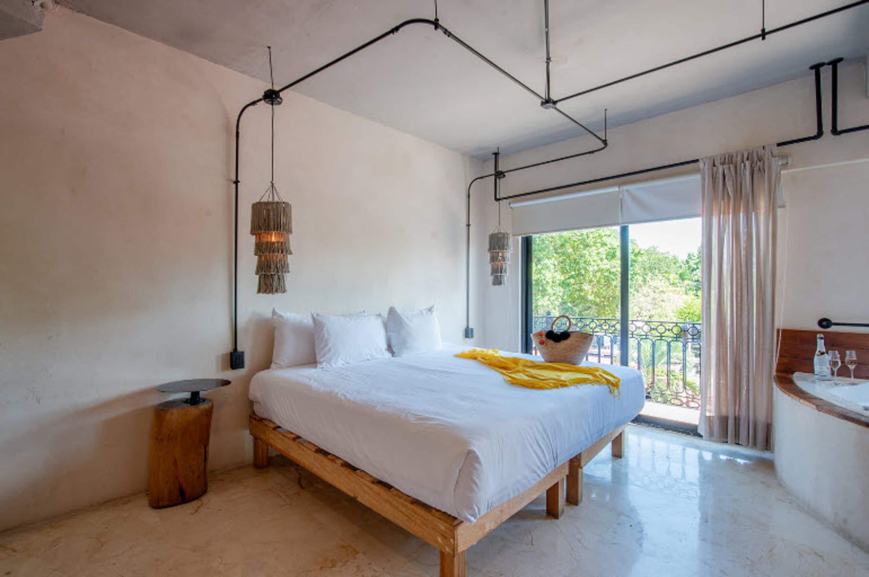 A bedroom with a large bed, a wooden nightstand, a hanging light fixture, and a view of the outdoors through a window.