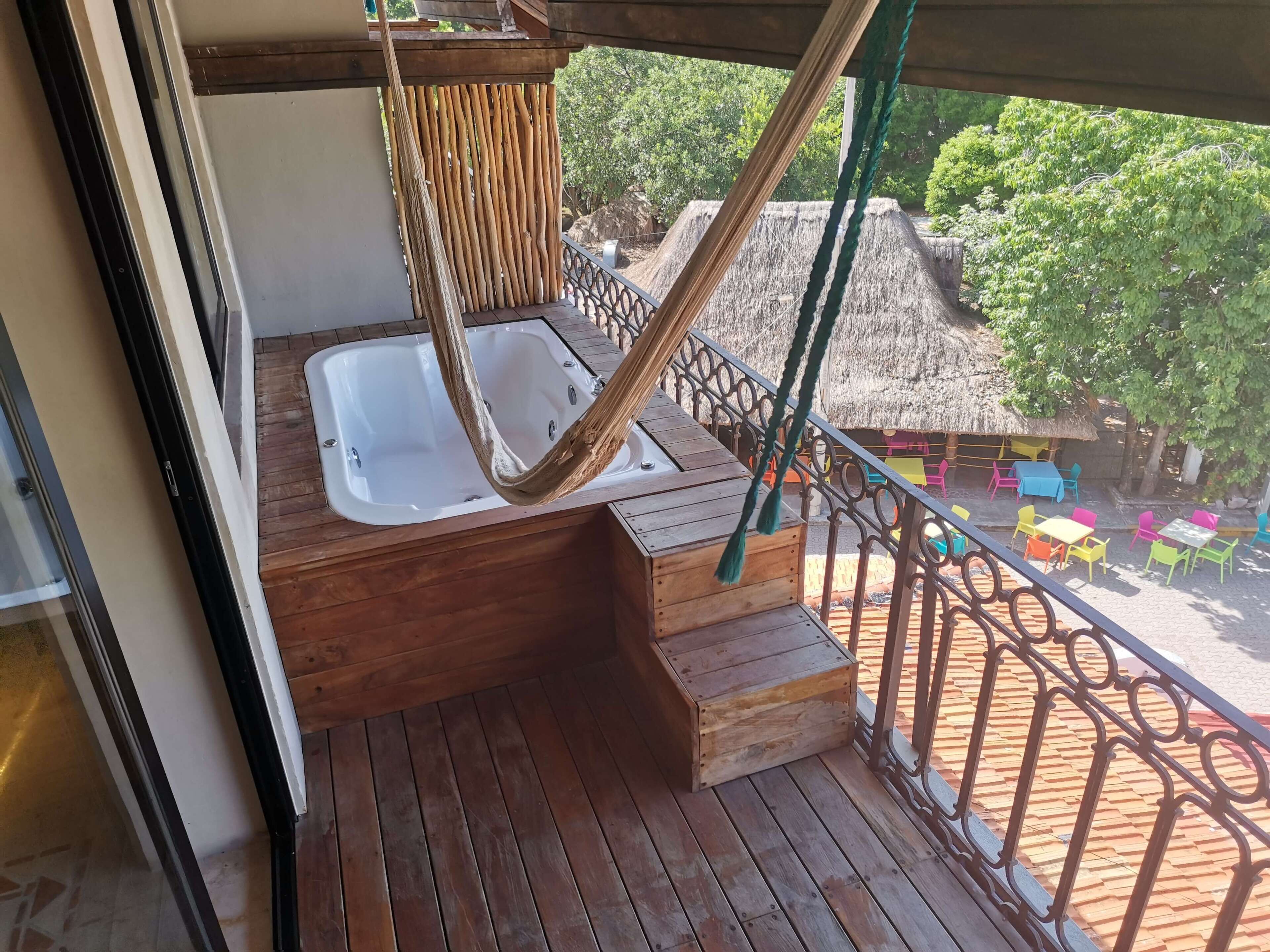 Una terraza de madera con un jacuzzi, una construcción con techo de paja y mobiliario exterior de colores.