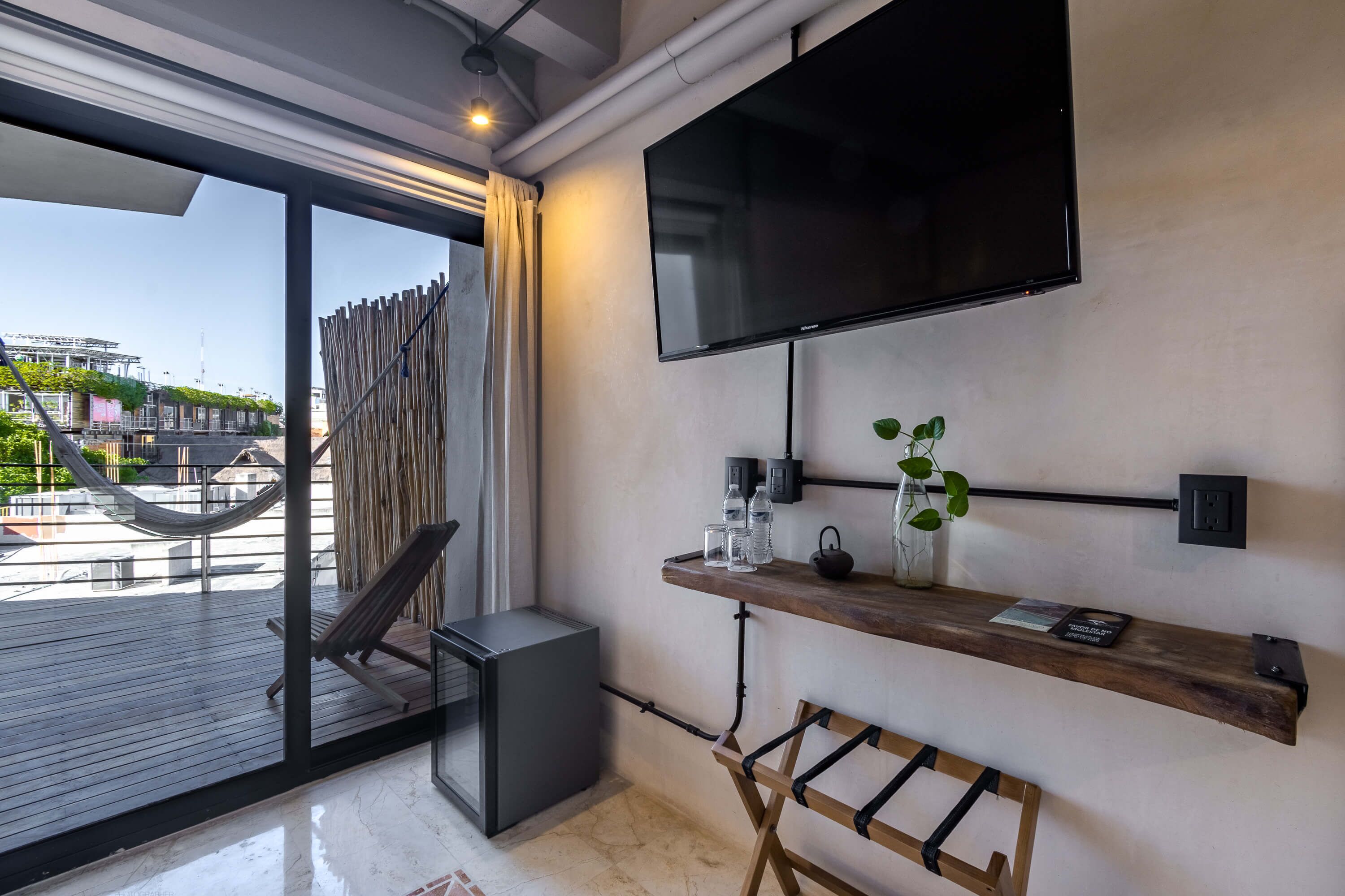 A modern room with a large TV, a wooden shelf with drinks, a hammock, and a view of a building outside.