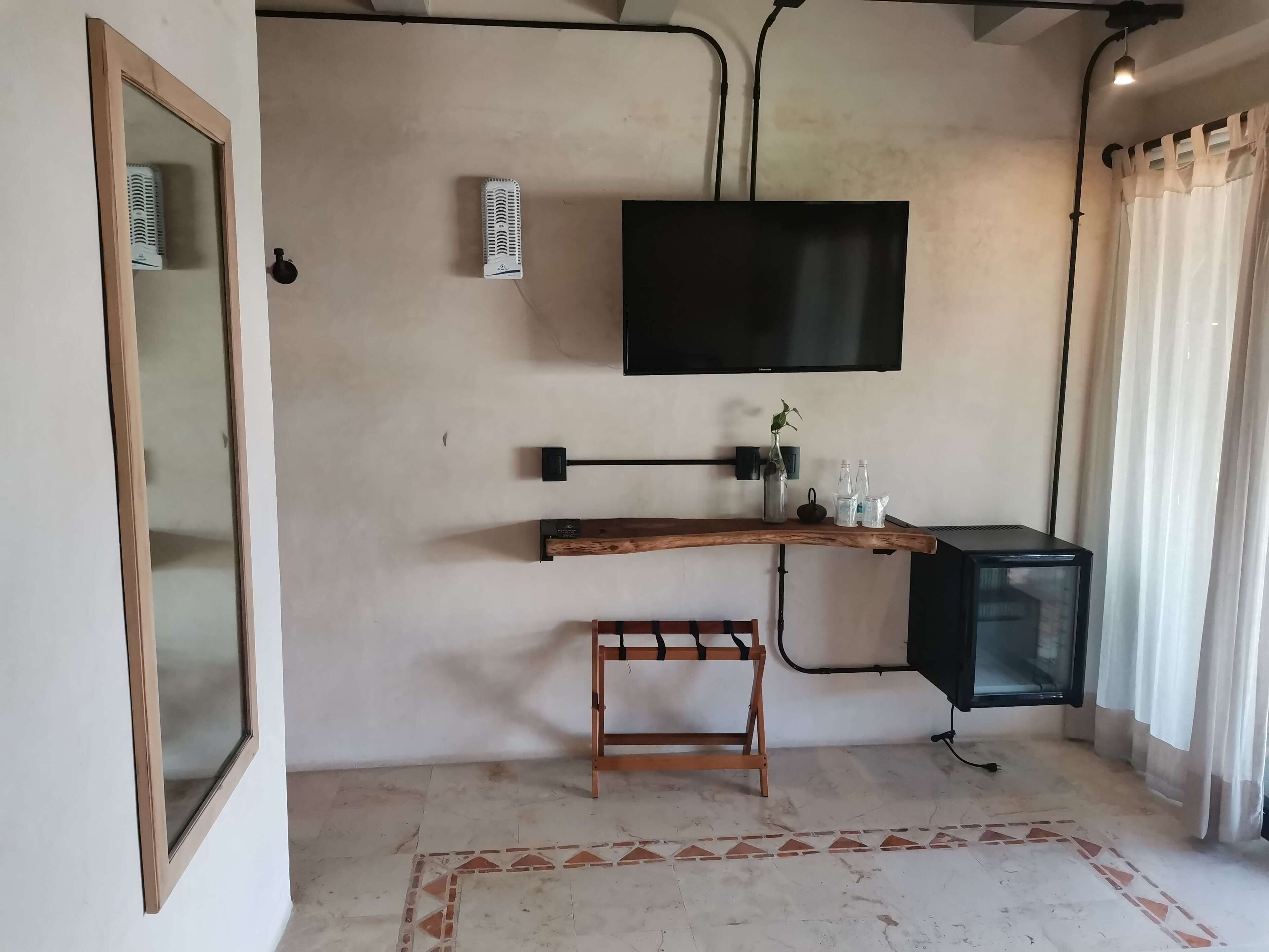 A room with a flat-screen TV mounted on the wall, a wooden desk with a chair, a small stool, and a cabinet with a TV.
