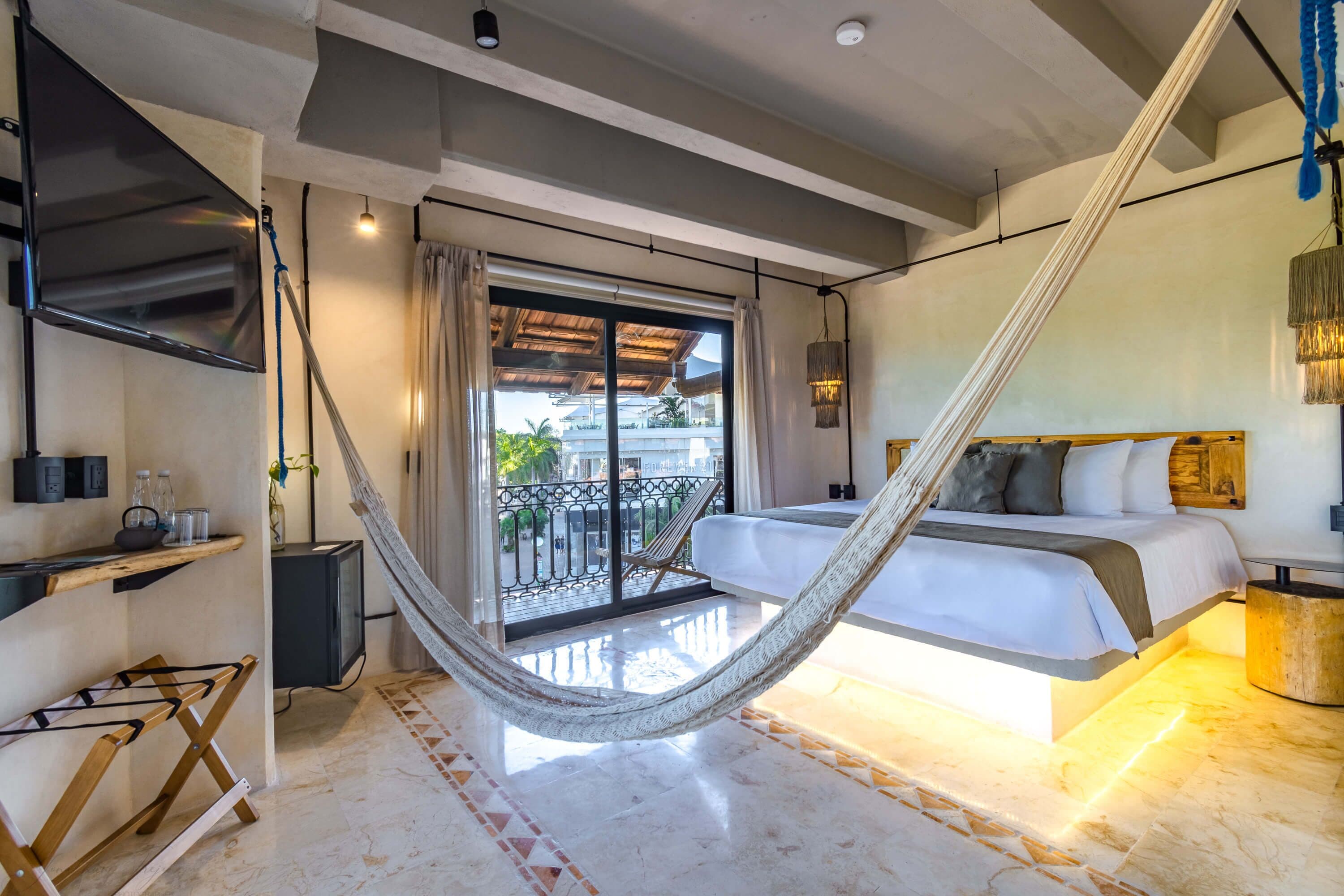 A modern hotel room with a large bed, a hammock, a balcony with a view, and a TV mounted on the wall.